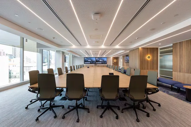 A long boardroom table with a light timber top, lined with dark office chairs, with a fluted timber wall, a round brass wall light and a blue upholstered bench at the far right.
