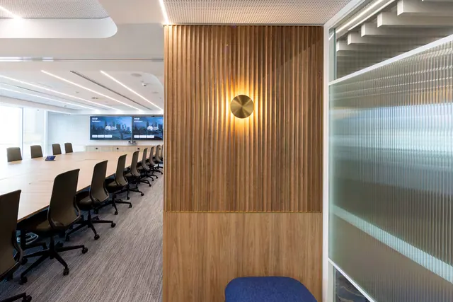 A fluted timber wall panel above a plain timber base, with a round brass wall light, next to a ribbed glass partition, with the boardroom table and chairs beyond.