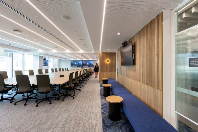 A long blue upholstered bench runs along a fluted timber wall in a boardroom, with round timber-topped side tables, a wall-mounted screen and the long boardroom table to the left.
