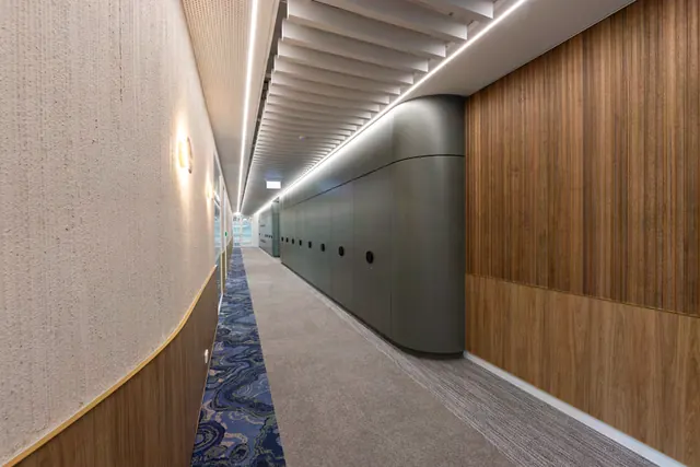 Tall green-grey storage cabinets with a rounded end and round black pulls line a corridor between a fluted timber wall and a curved wall with a timber lower panel.