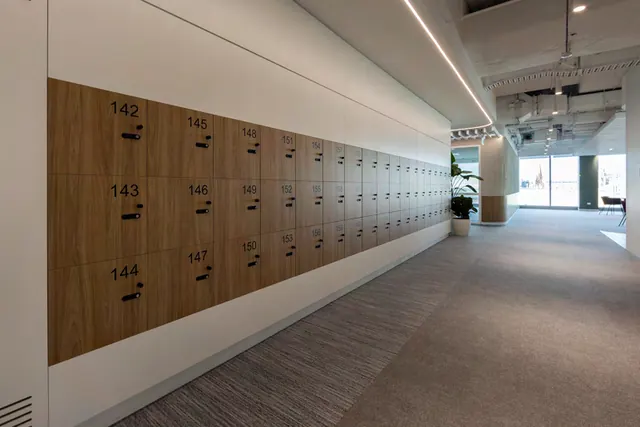 A long run of timber lockers with numbered doors and black locks, set into a white wall in an open-plan office.