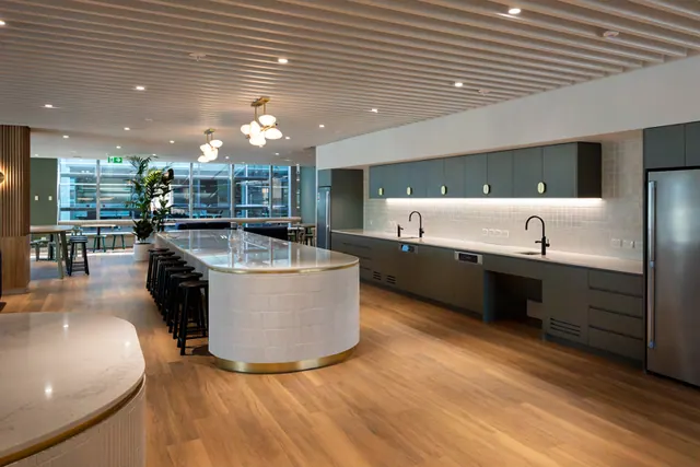A long kitchen island with a white tiled curved end, stone benchtop, brass base trim and black stools, facing a run of dark green cabinetry with overhead cupboards, a lit tiled splashback and a tall fridge.