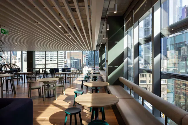 A long upholstered bench seat runs along floor-to-ceiling windows in a sunlit cafe area, with round timber tables, stools and a view over the city.