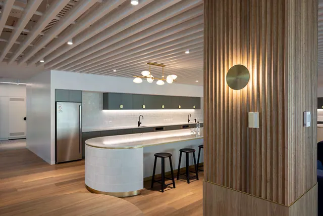 A kitchen with a white tiled island with a curved end, stone benchtop and black stools in front of dark green cabinetry and overhead cupboards, seen past a fluted timber-clad column with a round brass wall light.
