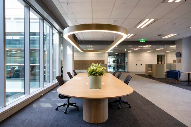 An oval meeting table with a light timber top on a round timber pedestal base, with a potted plant and black task chairs, under an oval pendant light beside floor-to-ceiling windows.