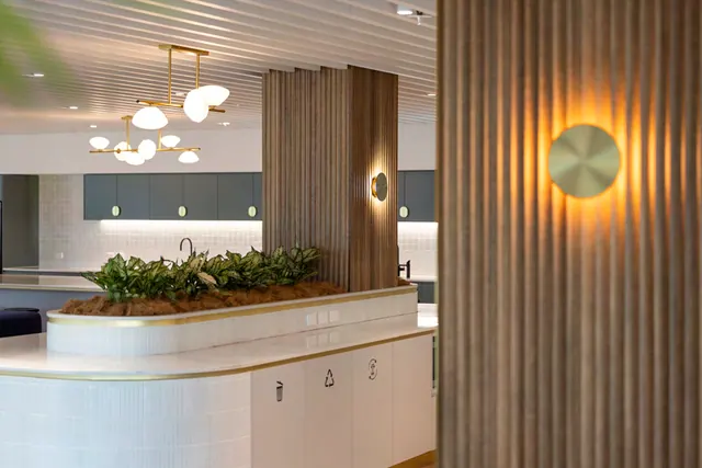 A white tiled island with an integrated planter, brass trim and white cabinet doors marked with waste and recycling symbols, beside a fluted timber column with a round brass wall light.
