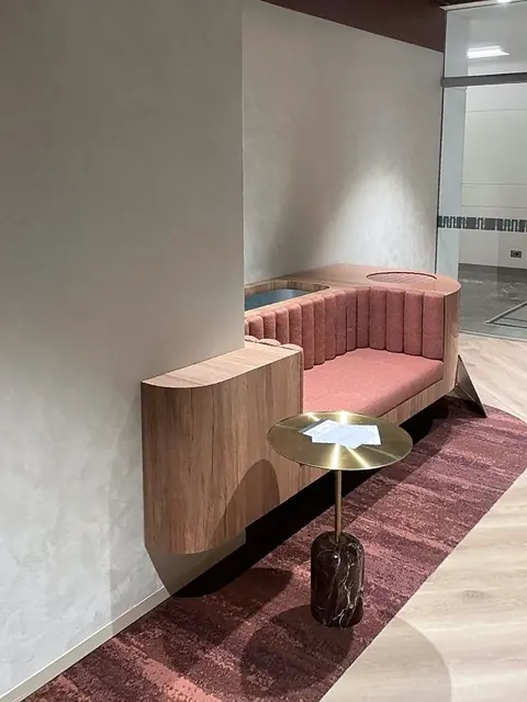 Coral fluted banquette with a rounded timber end and built-in planter, set into a wall recess beside a glass-walled corridor, with a brass side table in front.