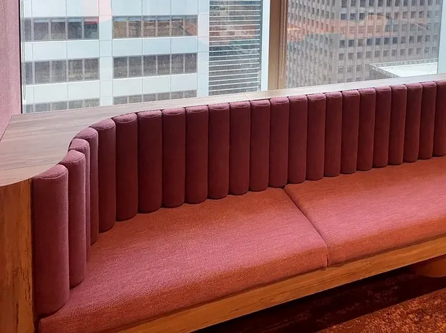 A long banquette with fluted raspberry upholstery, seat cushions and a timber base and end panel runs beneath a window overlooking city towers.