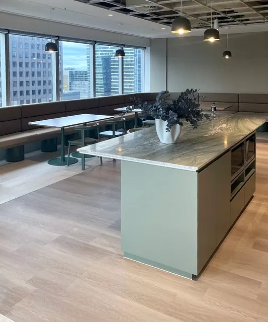 A sage-green island with a veined stone top and built-in appliances stands in a breakout space with upholstered banquette seating along the windows.
