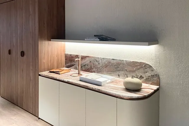 Coffee station with a pink-veined marble benchtop and curved splashback, curved off-white base cabinets, a lit floating shelf and a tall walnut cabinet.