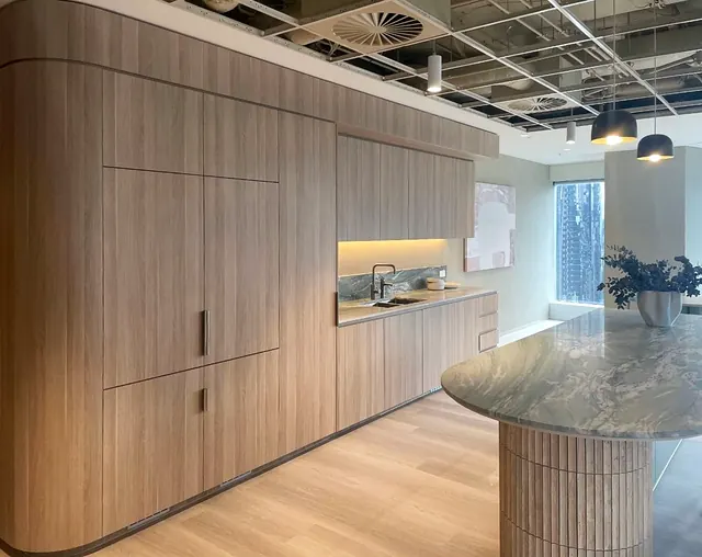 A run of tall light-oak cabinetry with a rounded end panel leads into a kitchen with overhead cupboards and a lit stone splashback, beside a stone-topped island on a fluted base.