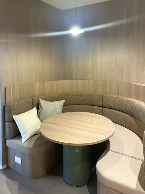 Curved booth seating in olive fabric around a round timber table on a green pedestal, set against curved timber-veneer wall panels under a pendant light.